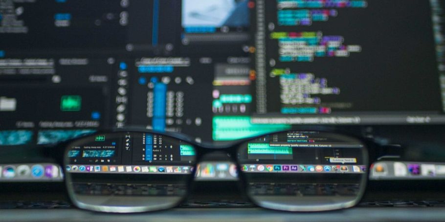 Brille auf Schreibtisch, im Hintergrund Monitore mit komplexen Daten